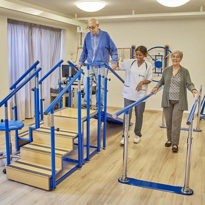 Dos persones grans a la sala de fisioteràpia fent exercicis amb la supervisió de la fisioterapeuta